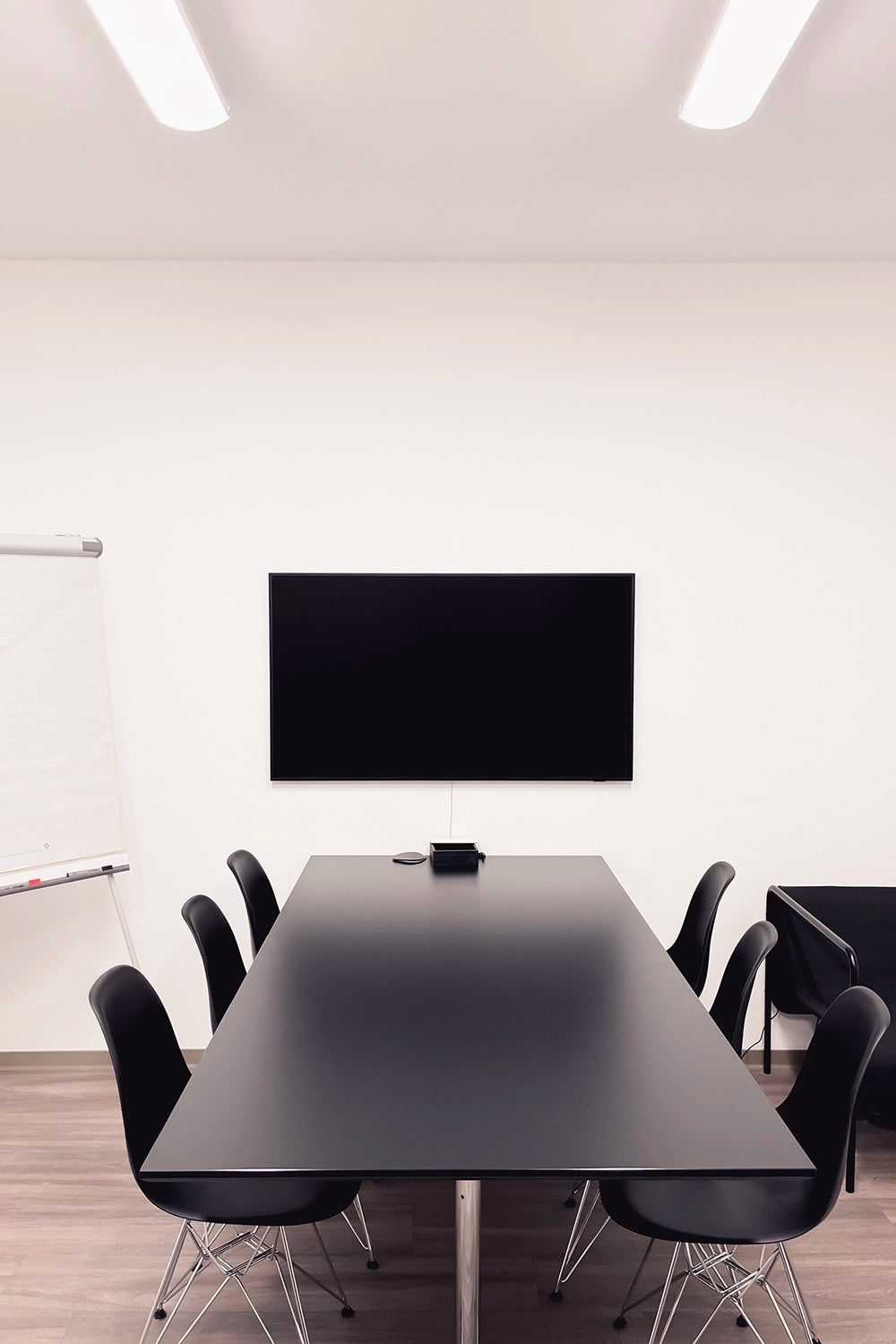 Konferenzraum mit schwarzem Tisch, Stühlen, einem Bildschirm und Flipchart.