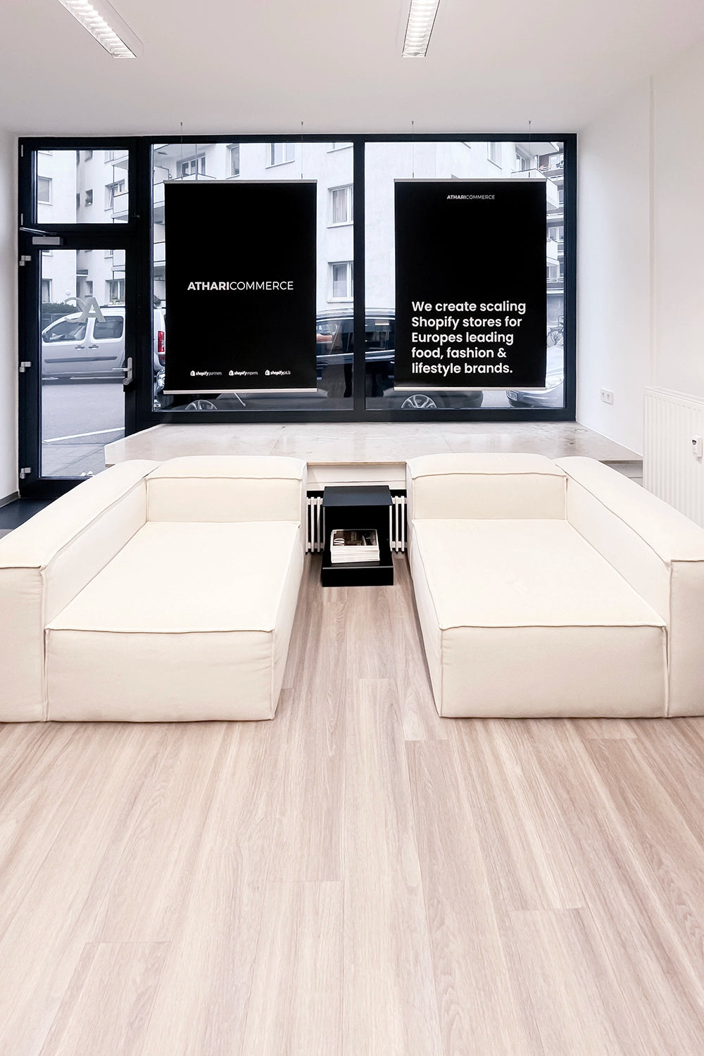 Moderner Raum mit beigen Sofas, Holzboden und Fenstern mit schwarzem Text: ATHARICOMMERCE.