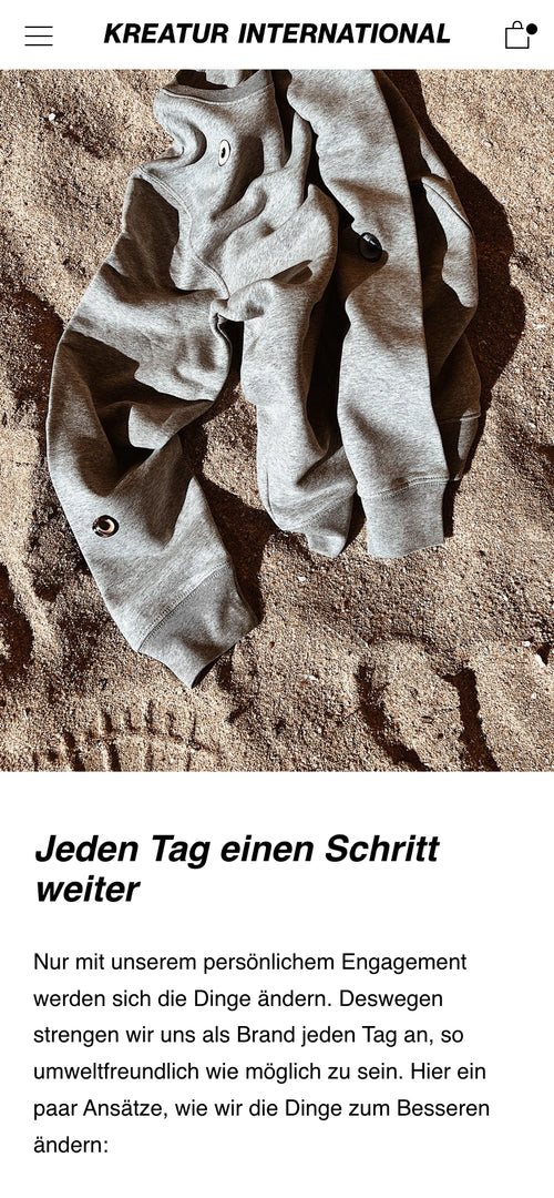 Graues Sweatshirt liegt auf Sand. Text darunter: Jeden Tag einen Schritt weiter.