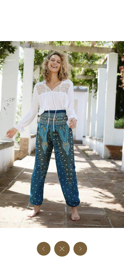 Frau in blauer Haremshose mit Muster und weißer Spitzenbluse, steht draußen und lächelt.